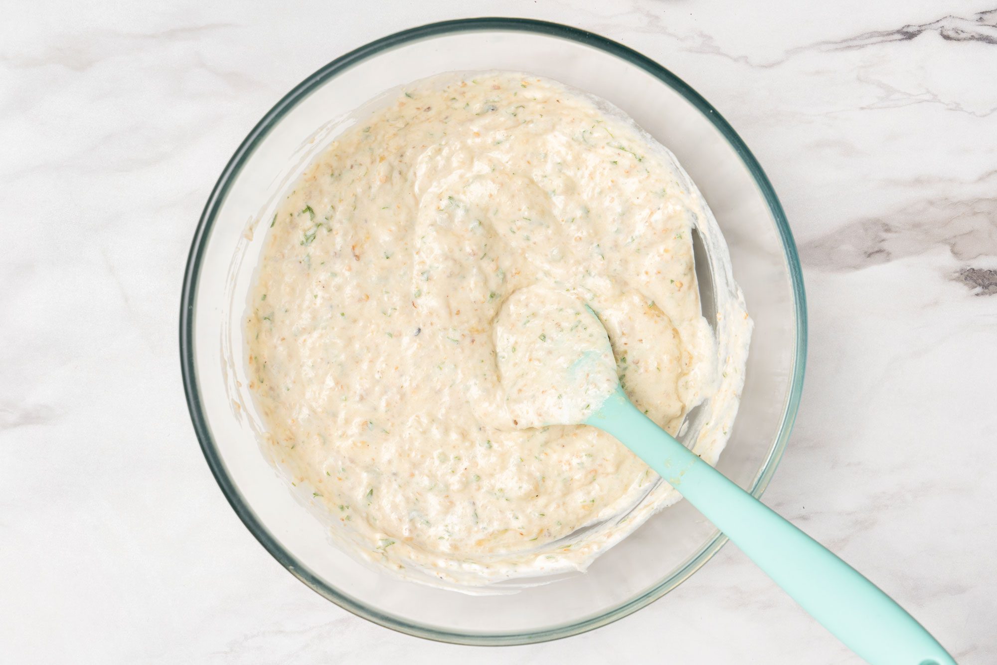Overhead shot of a glass bowl filled with a creamy, speckled mixture sits on a marble surface; A light blue spatula rests in the bowl, partially covered with the mixture;