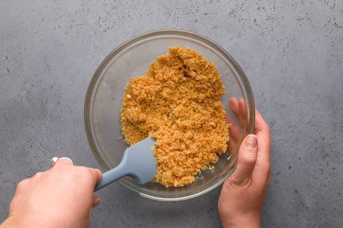 overhead shot of a person mixes a crumbly, golden brown mixture in a clear glass bowl with a gray spatula on a textured gray surface