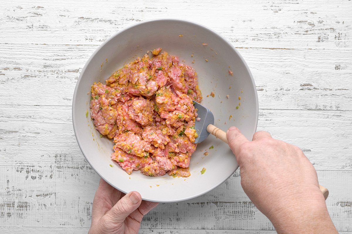 Two hands mix ground meat and seasonings in a gray bowl with a spatula on a white wooden surface.