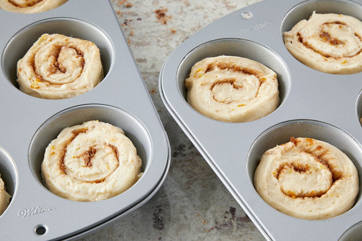Clsoe shot of unbaked rolls arranged in metal muffin tins; showing spiral swirls of cinnamon filling; the tins rest on a lightly speckled countertop.