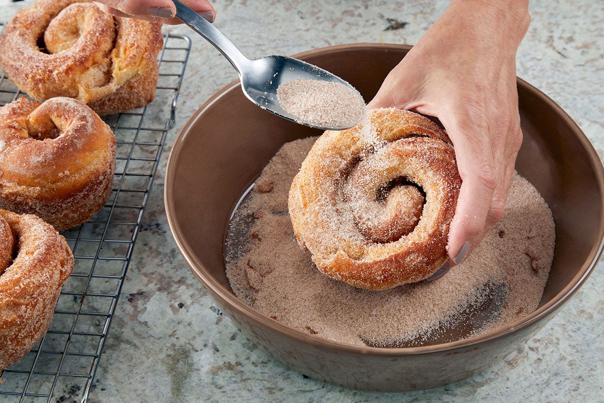 Close-up shot of a hand dipping a roll into a bowl of cinnamon sugar and sprinkling more on top with a spoon; while additional rolls cool on a wire rack nearby.