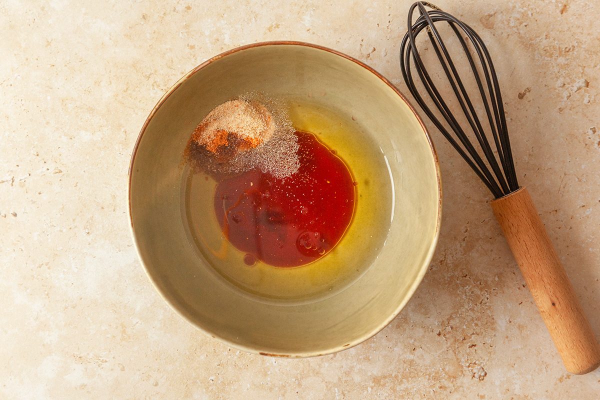 overhead shot of a green bowl containing oil, vinegar, and spices sits on a tan countertop next to a metal whisk with a wooden handle
