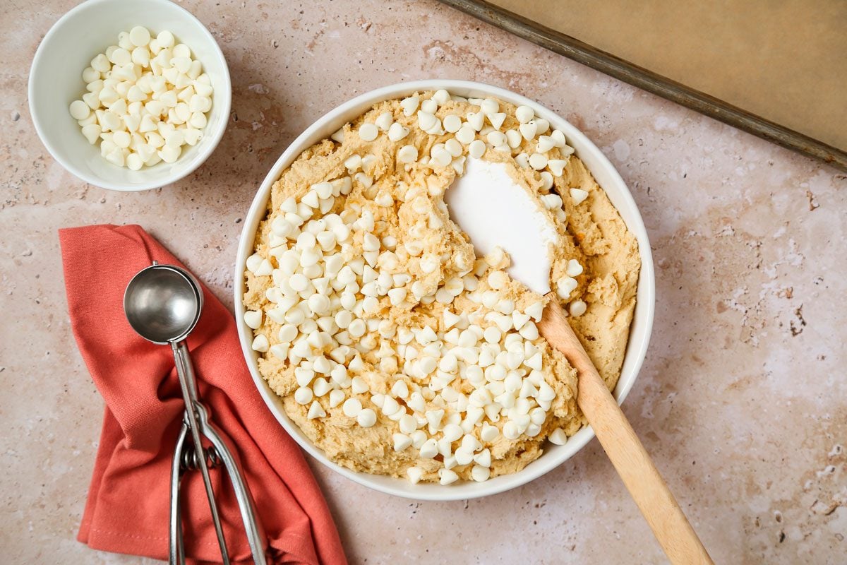 Overhead shot of a large bowl cream butter and sugars until light and fluffy 5-7 minutes; Beat in eggs, orange zest and extracts; In another bowl whisk flour, pudding mix and baking soda; gradually beat into creamed mixture; Stir in white baking chips; spatula; a scoop spoon over a orange napkin; all set on a texture marble surface;