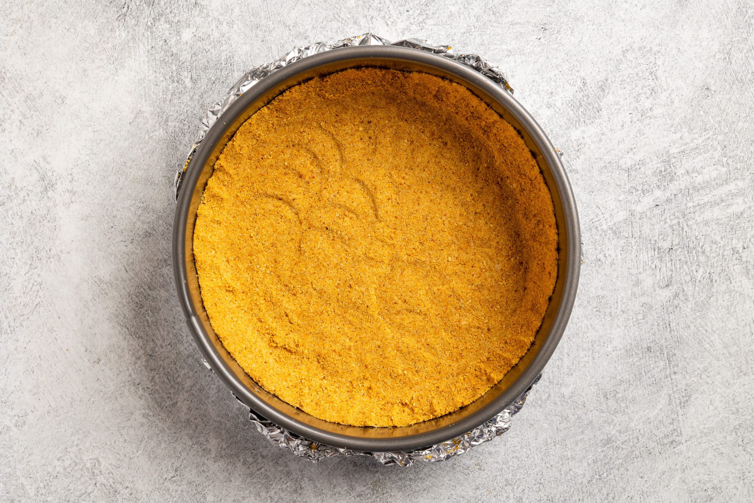 A round baking pan lined with foil holds a pressed graham cracker crust, ready for filling, on a light gray textured surface.