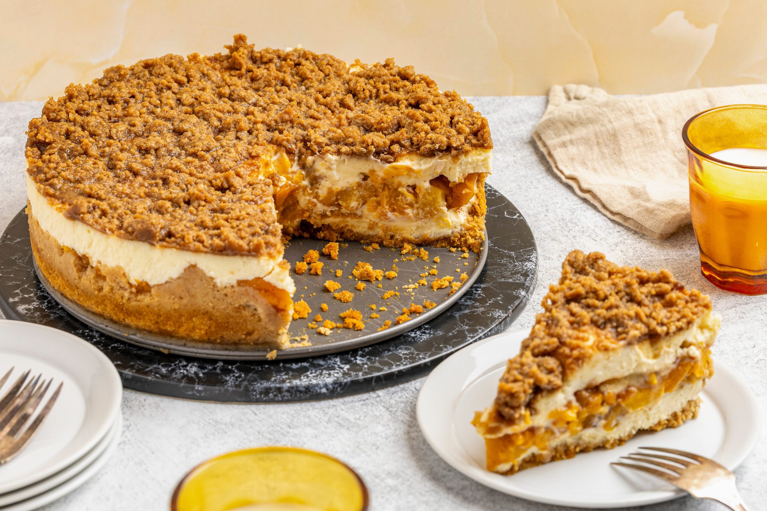 A crumb-topped pie with a large slice removed sits on a black platter. The pie has a creamy filling and visible fruit layer, with a slice served on a plate nearby. Forks and yellow glasses are on the table.