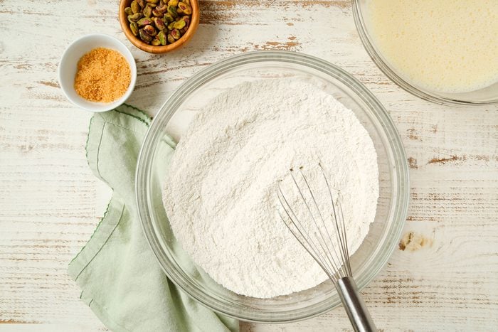 overhead shot of a glass bowl filled with flour and a whisk sits on a white wooden surface, Nearby are a green cloth, a small bowl of brown sugar, a bowl of pistachios, and a bowl of liquid batter