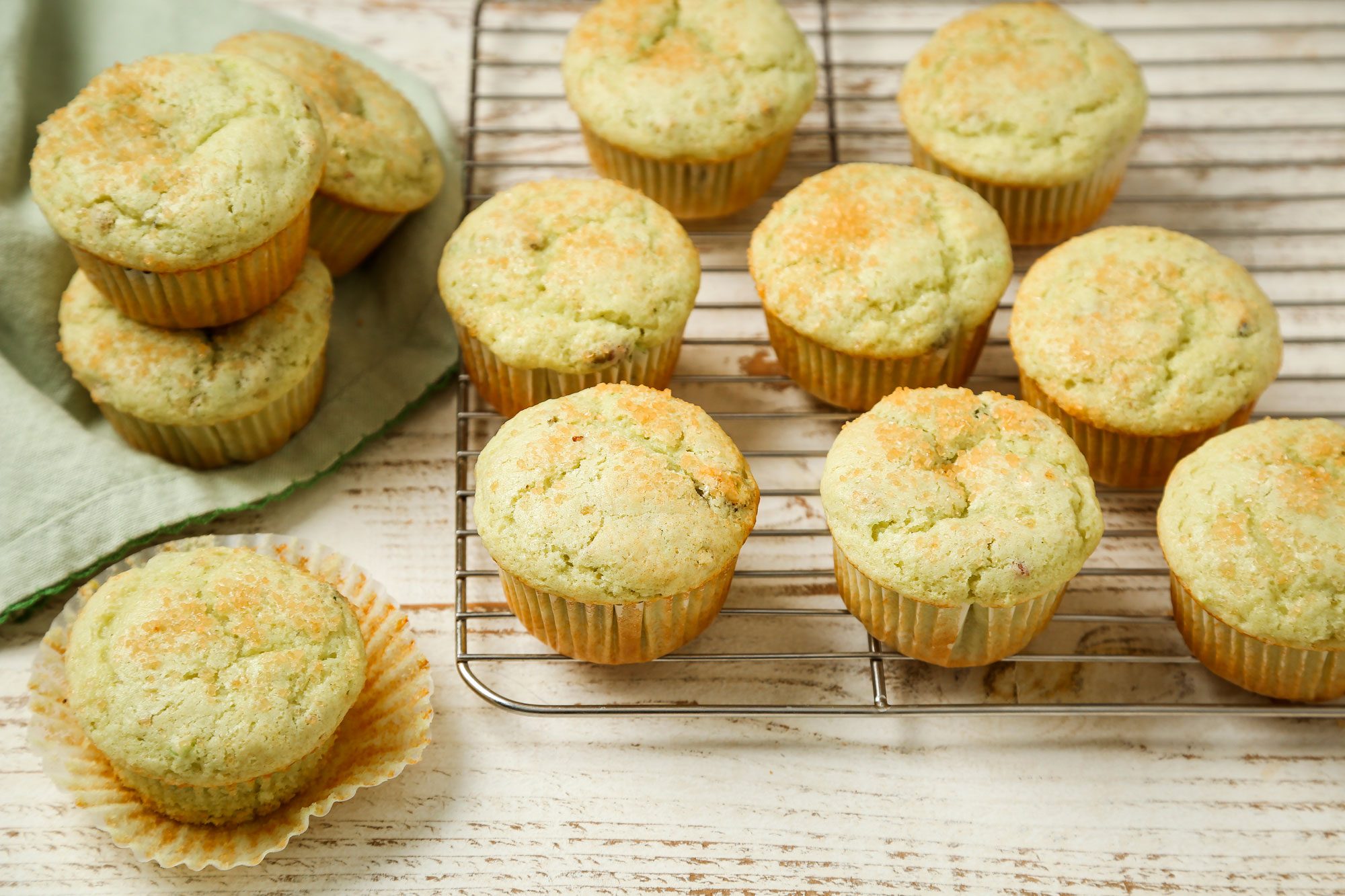3/4th shot of a batch of freshly baked Pistachio muffins cooling on a wire rack and a rustic white wooden surface, with a few muffins placed beside on a green cloth and one muffin partially unwrapped in the foreground
