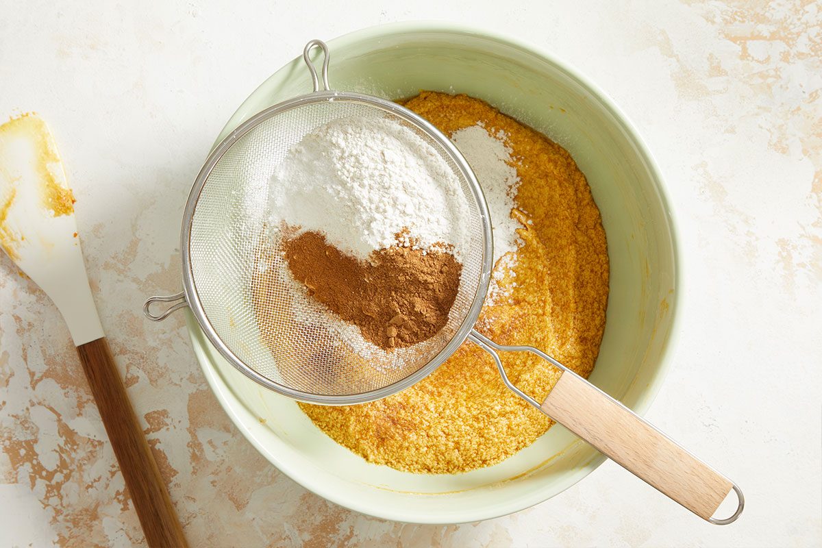 A mixing bowl with cornmeal and other ingredients, topped with a mesh sieve holding flour, baking powder, and cinnamon; a spatula rests nearby on a light countertop.