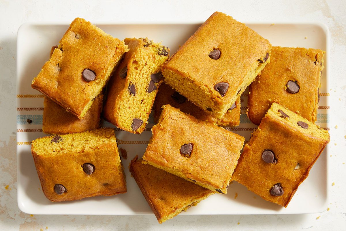 A rectangular white plate holds nine golden-brown squares of pumpkin chocolate chip bars, each bar featuring visible chocolate chips on top. The bars are arranged in a scattered fashion.