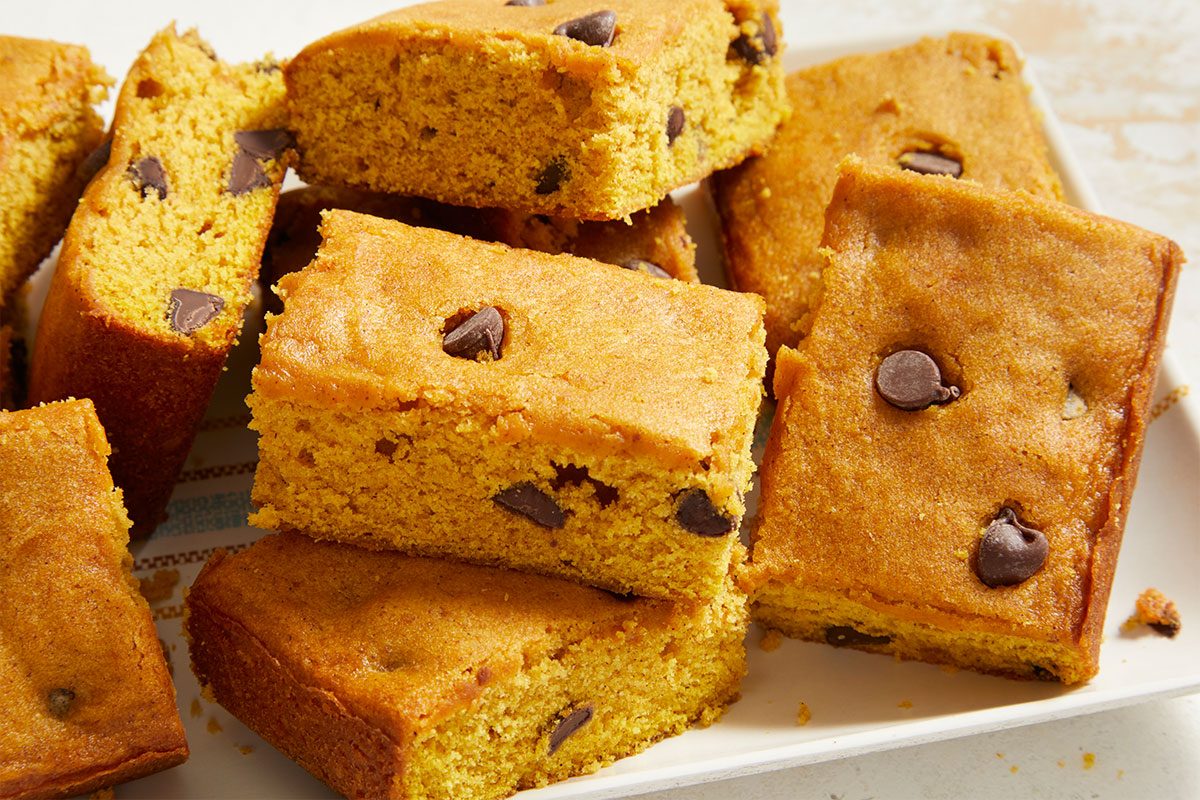 Several rectangular pumpkin chocolate chip bars are stacked on a white plate. The bars are golden brown and studded with chocolate chips.