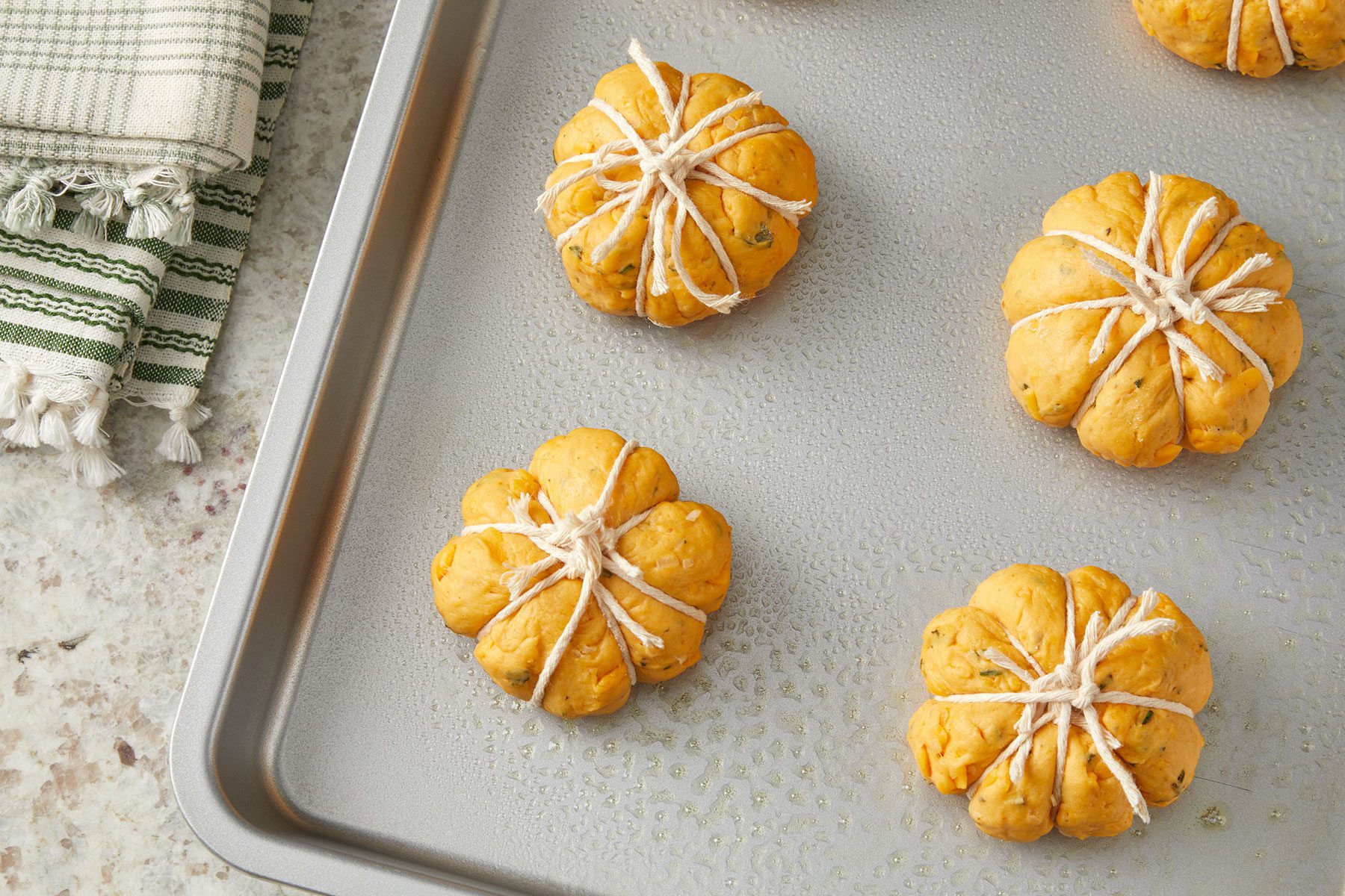 Wrapped dough balls in kitchen strings placed on a greased baking dish with a kitchen towel on side.