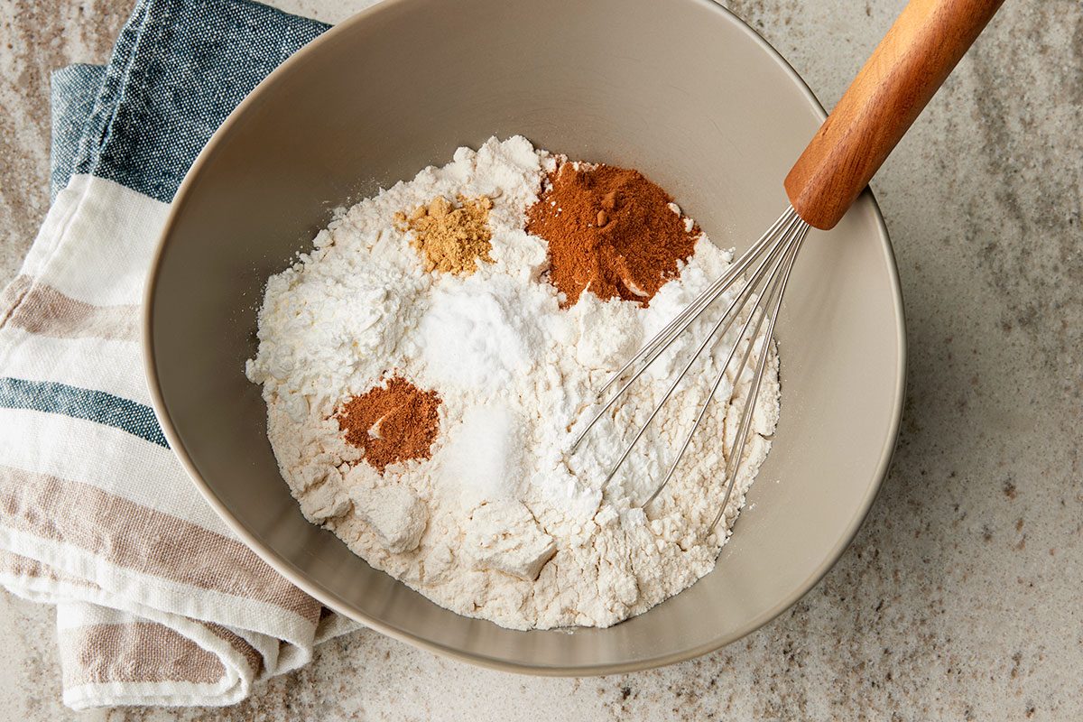 Overhead shot of a mixing bowl with flour, baking powder, and spices including cinnamon and ginger; placed beside a whisk and a striped kitchen towel on a countertop;