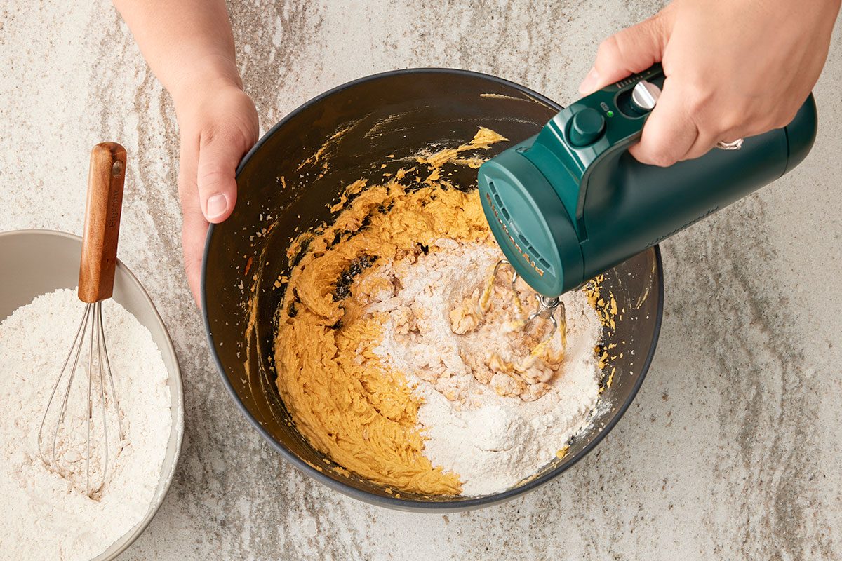 Overhead shot of a person using a green electric hand mixer to blend flour into a bowl of cookie dough; a bowl of dry ingredients with a whisk rests nearby on a light countertop;