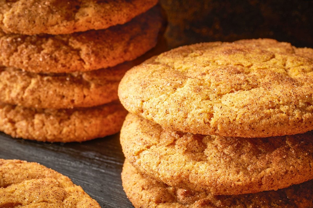 Close-up shot of pumpkin snickerdoodles, stacked and scattered on a dark slate surface; with golden-brown tops coated in cinnamon sugar;
