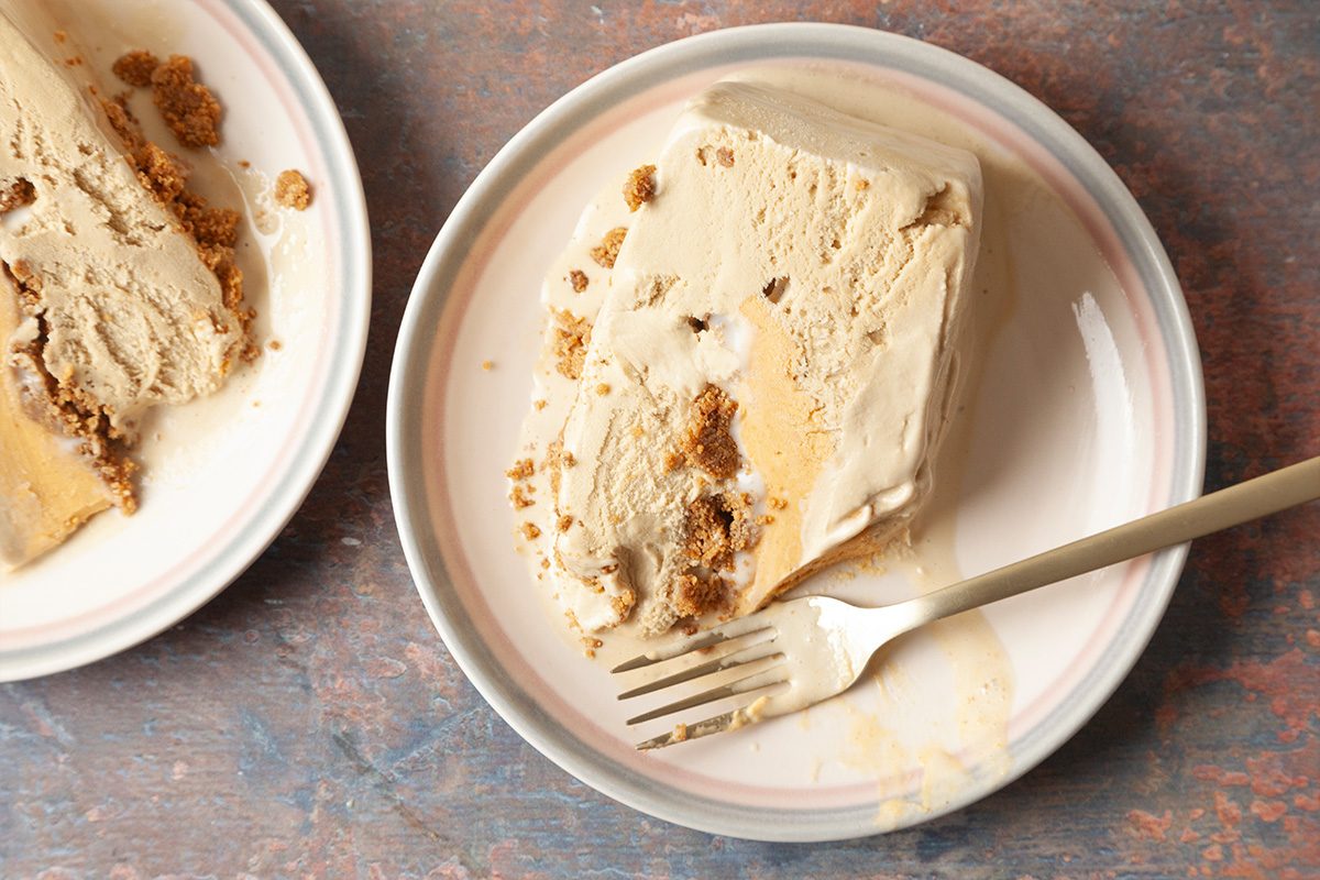 A slice of creamy, light brown pie with a crumbly crust on a white plate with a fork, set on a textured brown surface; another plate with pie is partially visible.