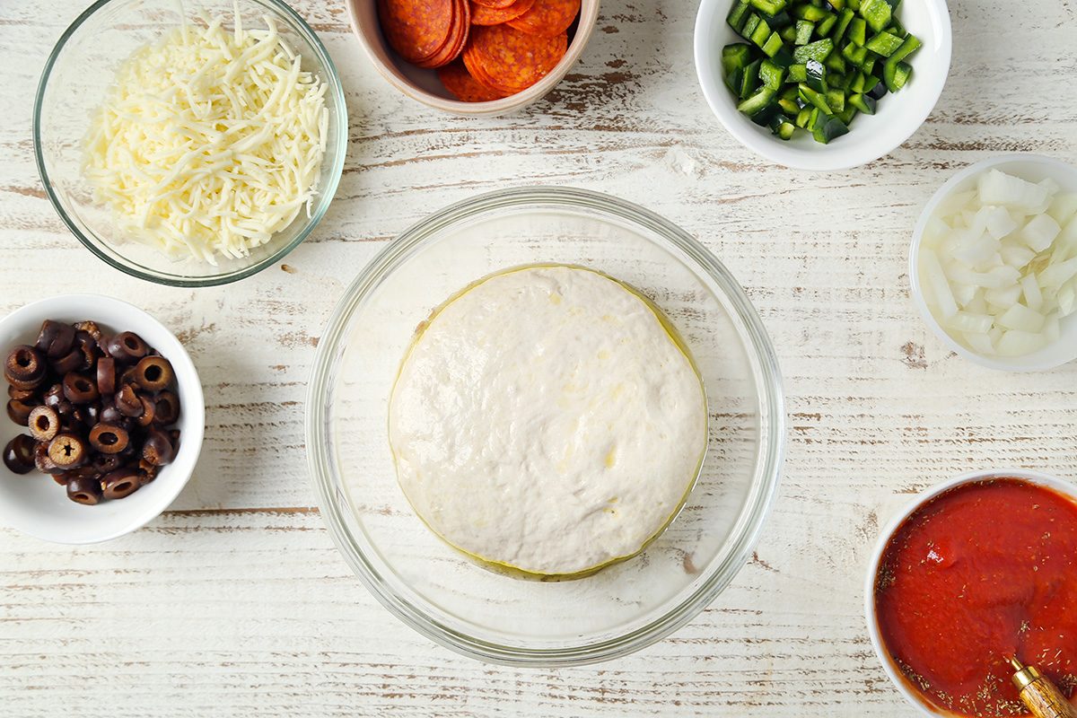 A bowl of pizza dough is surrounded by bowls of shredded cheese, pepperoni slices, chopped green peppers, chopped onions, sliced black olives, and tomato sauce on a white wooden surface.