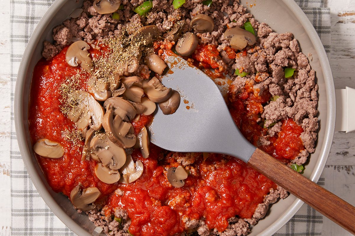 A skillet contains ground beef, tomato sauce, sliced mushrooms, chopped green peppers, and herbs, being mixed with a gray spatula. The skillet rests on a plaid cloth.