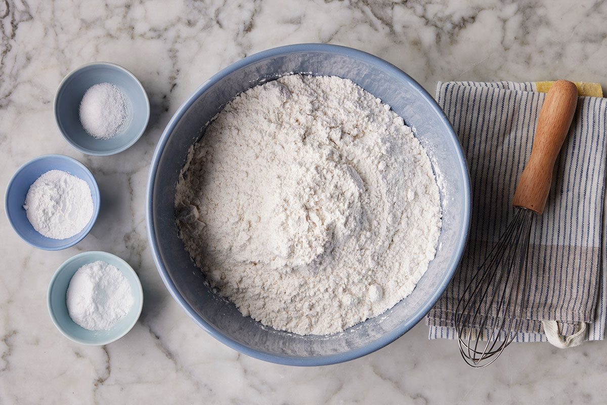 cake flour, baking soda, baking powder and salt are placed on a table in seperate bowls and ready to mix in the large bowl