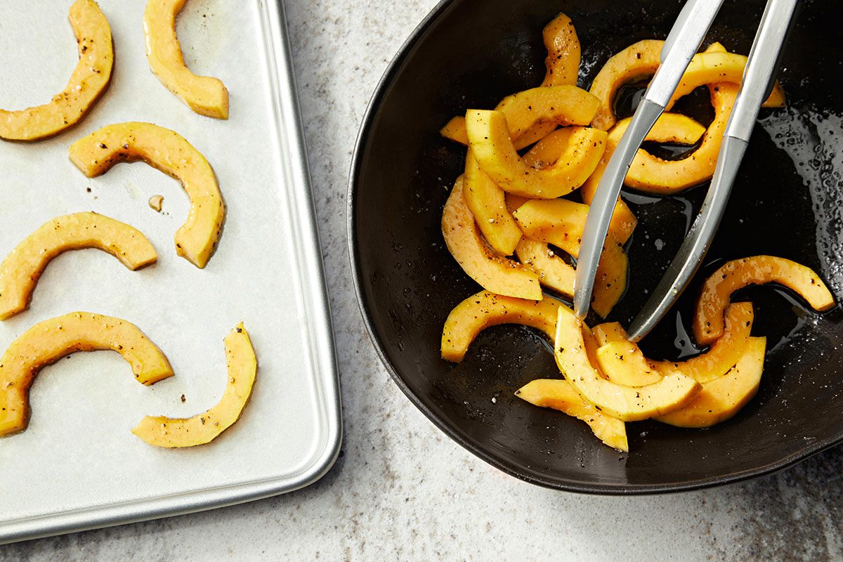 Overhead shot of sliced squash tossed with seasoning in a dark bowl with metal tongs; additional slices are neatly arranged on a baking sheet; ready for roasting.