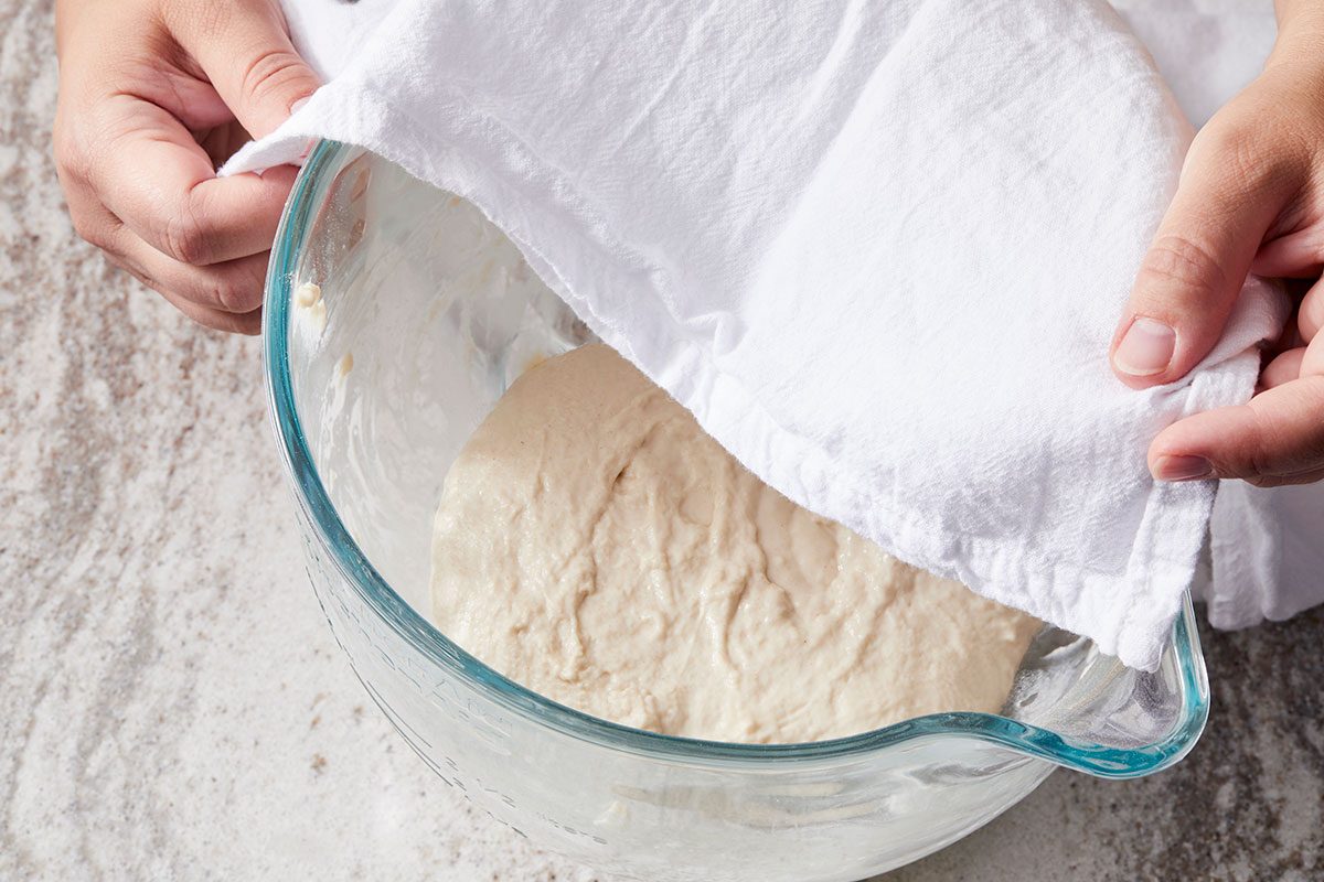 Overhead shot of a person lifting a white cloth to reveal rising dough in a glass mixing bowl on a light-colored surface.