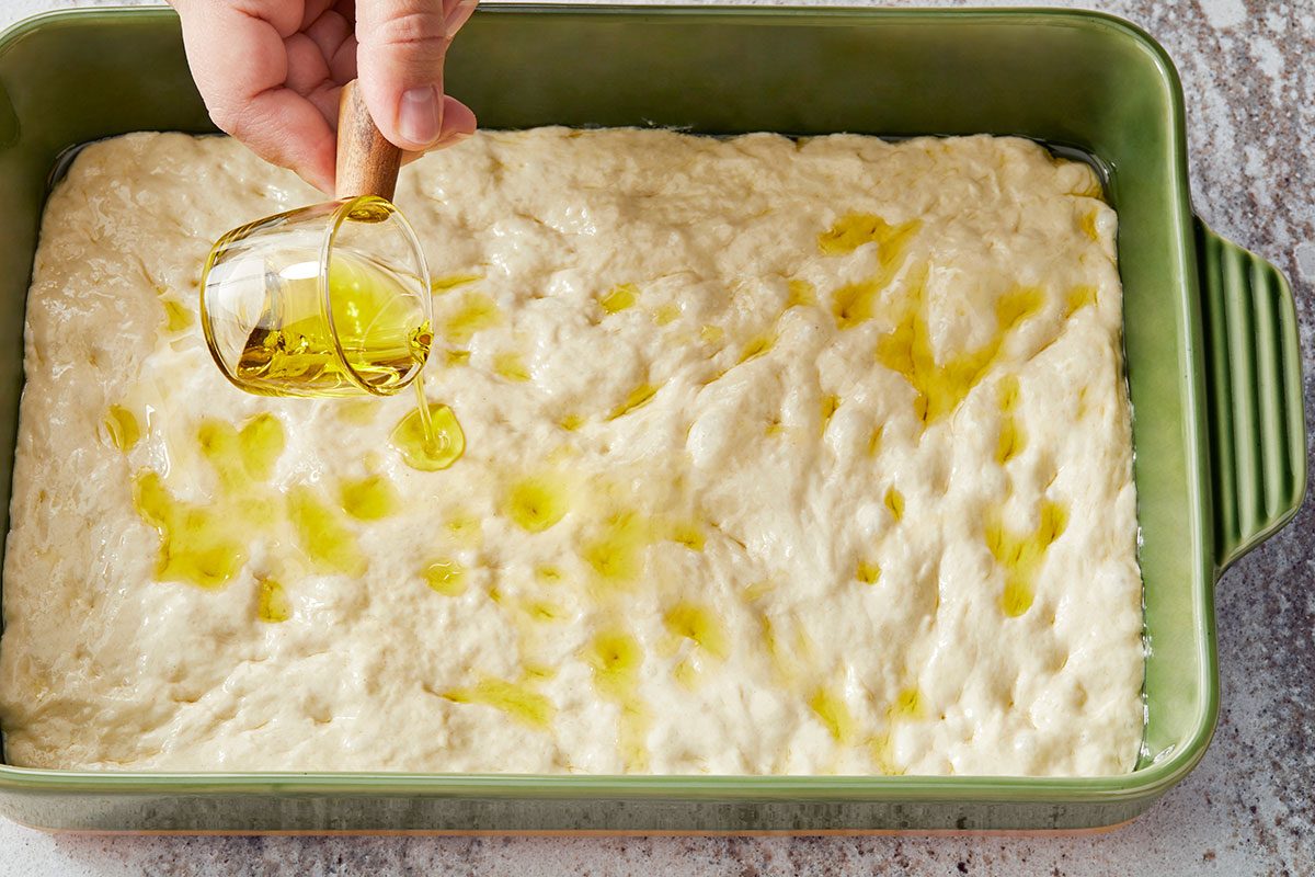 High-angle shot of a hand pouring olive oil from a small glass measuring cup over focaccia dough spread in a green rectangular baking dish.