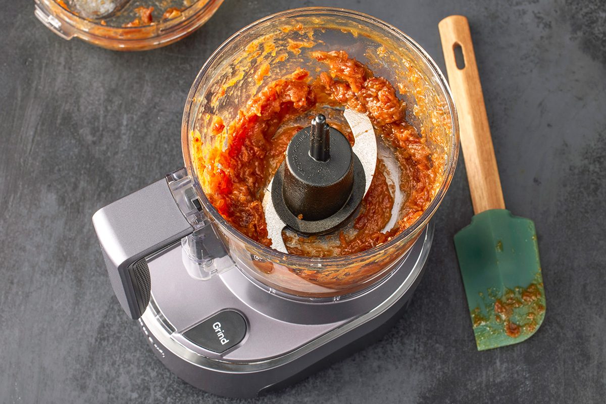 Overhead shot of a food processor filled with reddish sauce on a dark surface; with its lid set aside and a green spatula with sauce residue resting nearby
