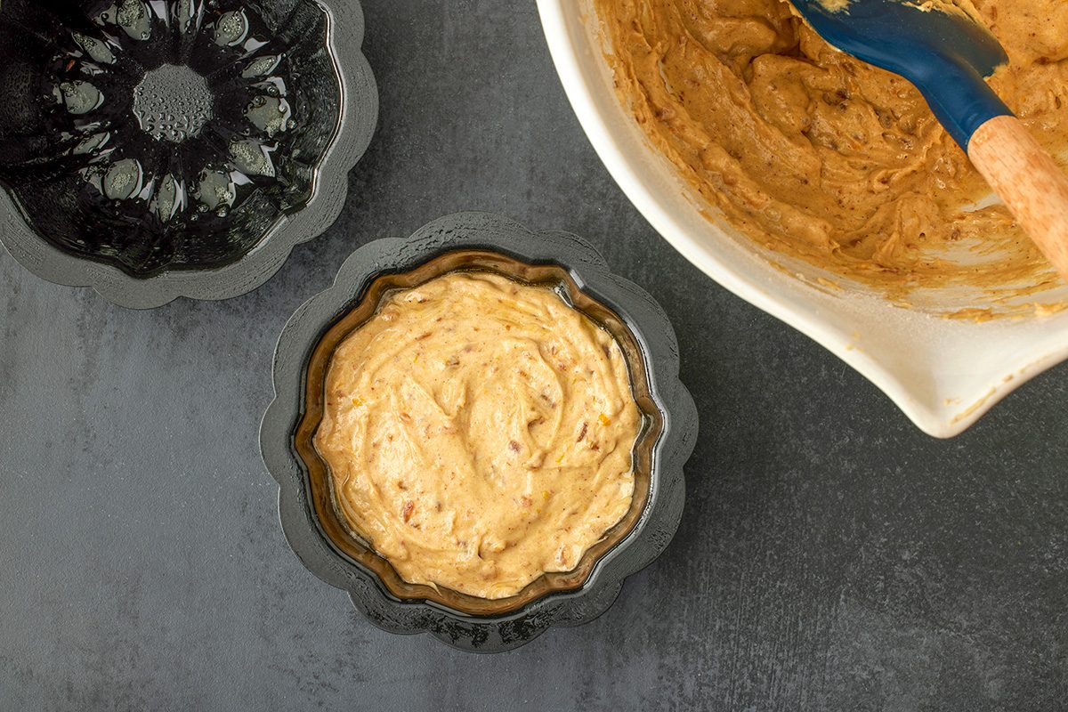 Overhead shot of a bundt cake pan filled with cake batter placed beside an empty greased pan and a mixing bowl with leftover batter and a spatula; all set on a dark countertop