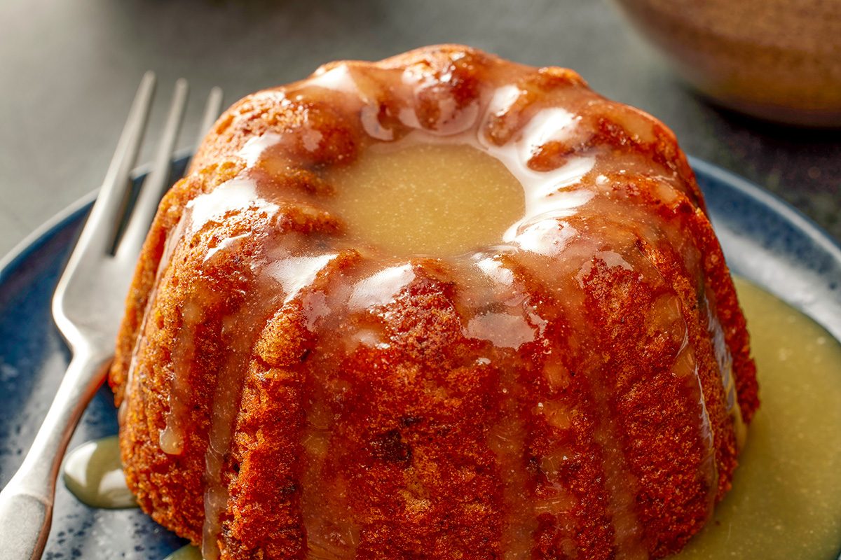 Close-up shot of a small round bundt cake drizzled with glossy glaze; placed on a dark plate with a fork beside it