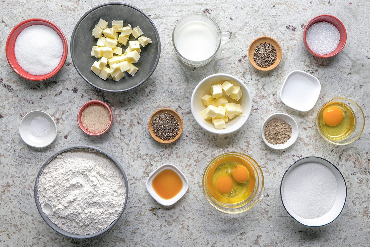overhead shot of Various baking ingredients are arranged on a stone countertop, including bowls of flour, sugar, butter cubes, milk, eggs, yeast, salt, vanilla extract, and measured spices