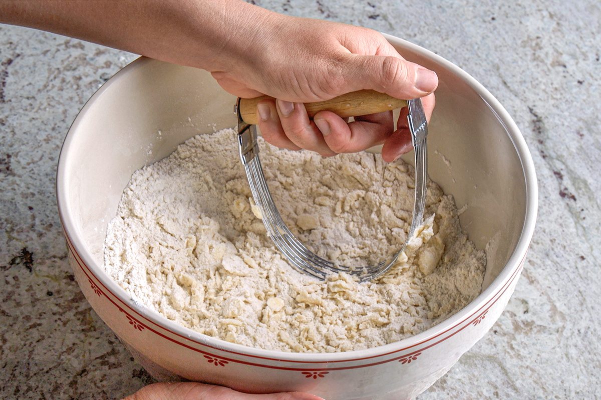 3/4th shot of A hand uses a pastry cutter to mix flour and butter in a large bowl; The bowl sits on a light, textured surface