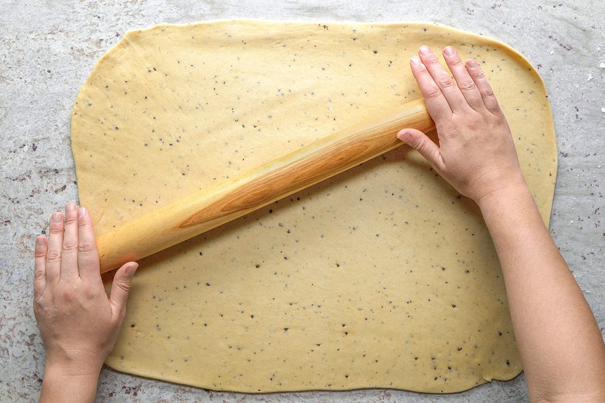 overhead shot of Hands rolling out speckled dough with a wooden rolling pin on a lightly floured, gray countertop