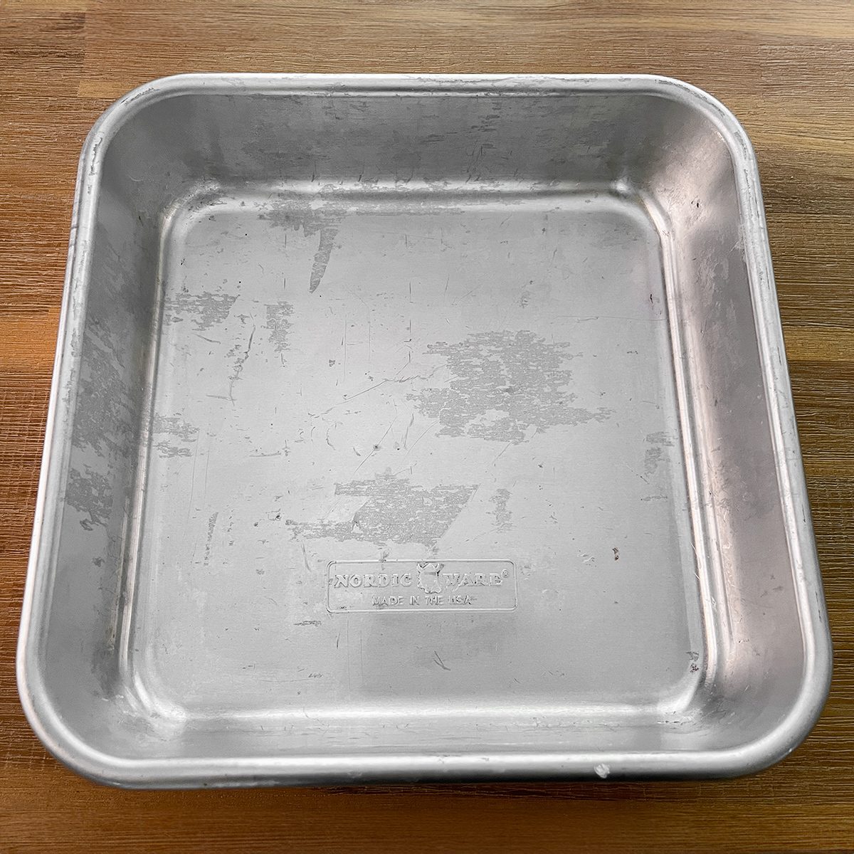 A square, metal baking pan with slightly worn surfaces sits on a wooden countertop.
