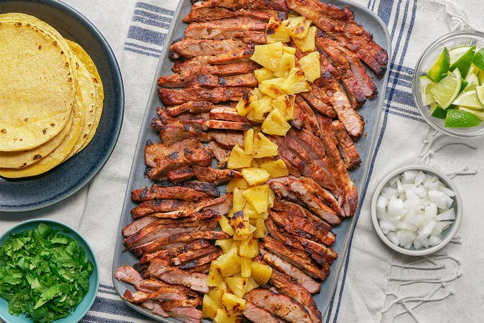 A platter of sliced, grilled meat topped with pineapple chunks, surrounded by bowls of chopped onions, cilantro, lime wedges, and a plate of corn tortillas on a striped cloth.