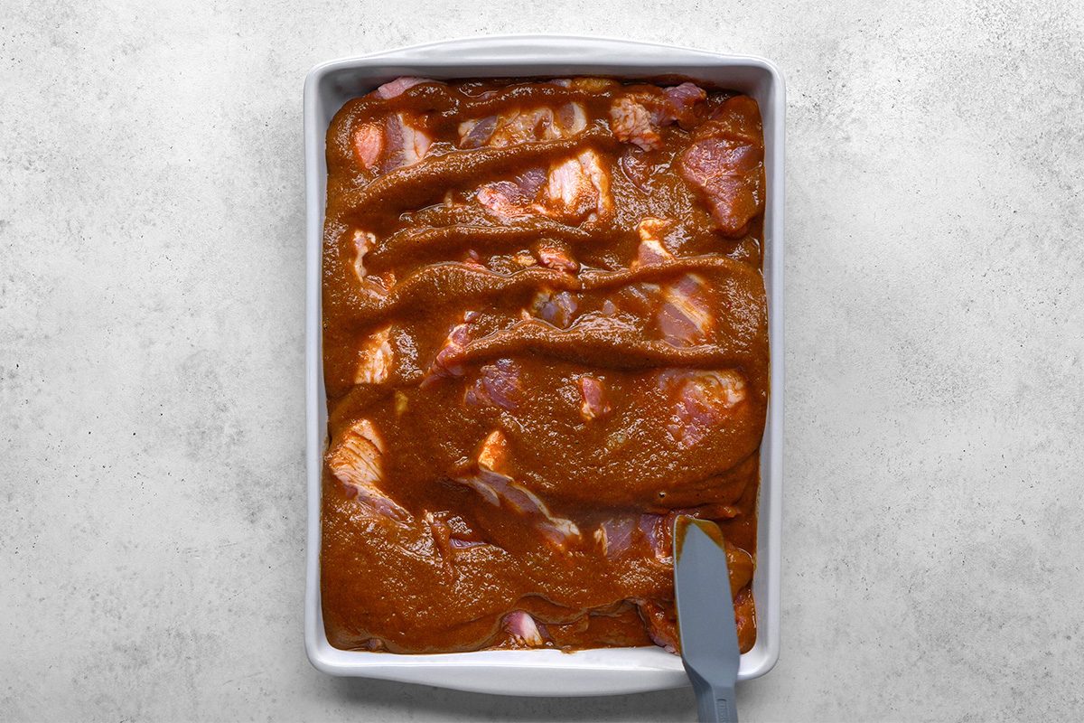 A rectangular white baking dish filled with raw meat pieces coated in a thick brown marinade, with a spatula resting in the bottom right corner of the dish.