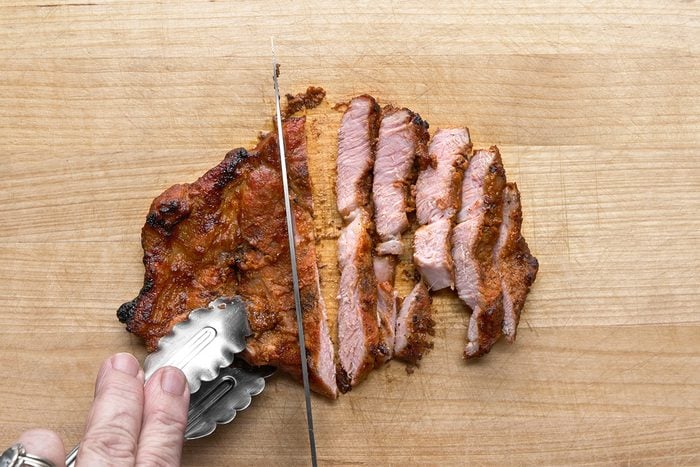A hand holds metal tongs gripping a piece of cooked, seasoned meat while a knife slices it into strips on a wooden cutting board.