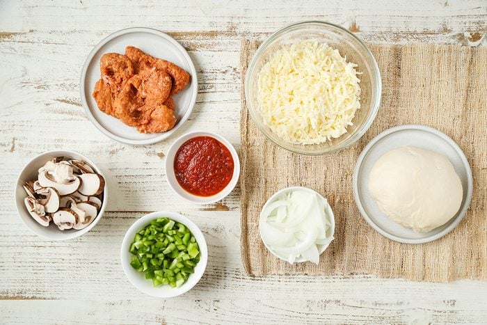 A top-down view of bowls containing pizza ingredients: shredded cheese, pizza dough, tomato sauce, green peppers, sliced mushrooms, seasoned chicken pieces, and a bowl of white sauce, all on a rustic white surface.