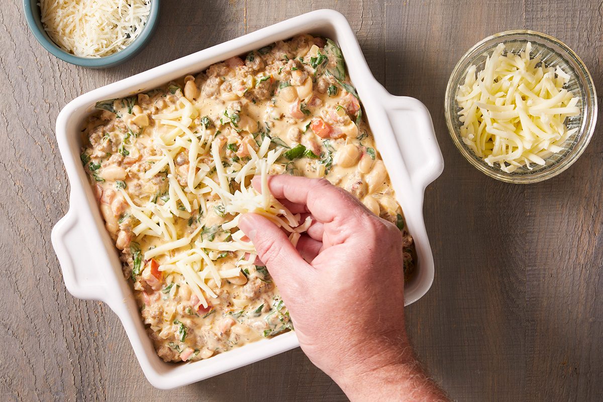 A hand sprinkles shredded cheese over a casserole in a square white baking dish, with bowls of cheese visible nearby on a wooden surface.