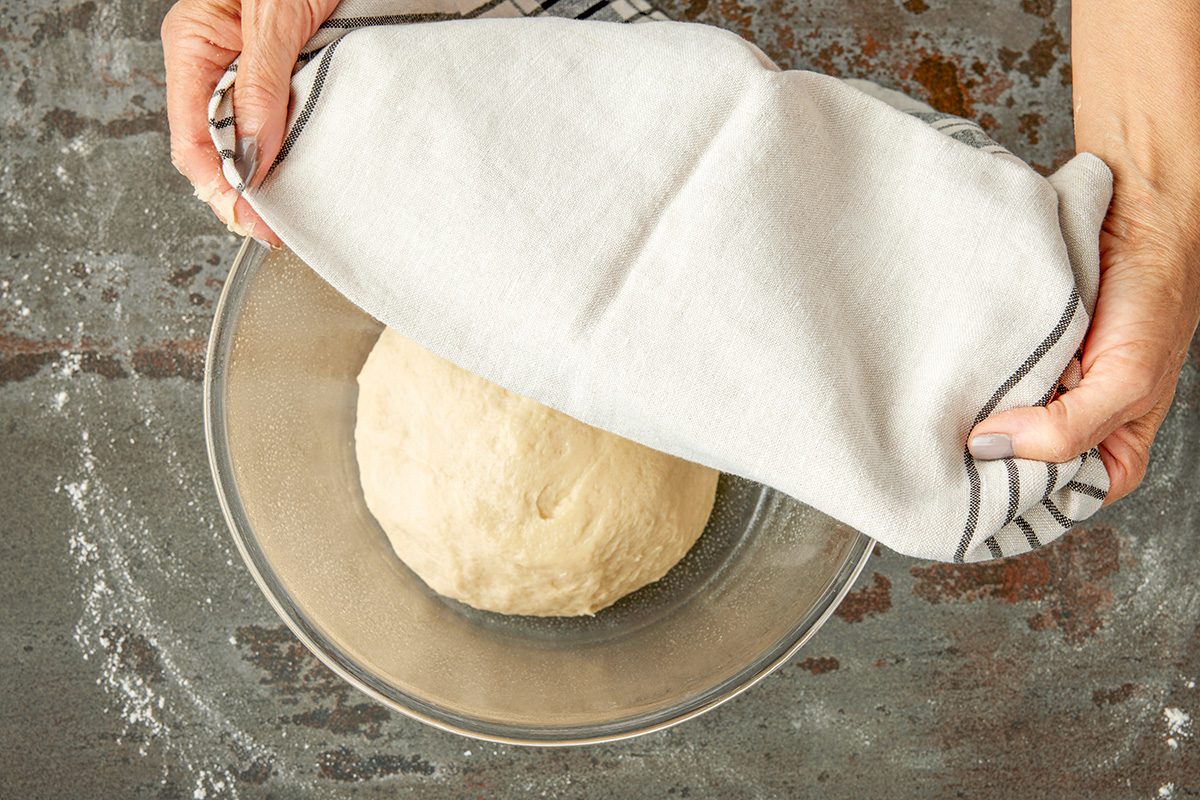 Overhead shot of a person covering a bowl of risen dough with a striped kitchen towel on a floured countertop; preparing it for proofing