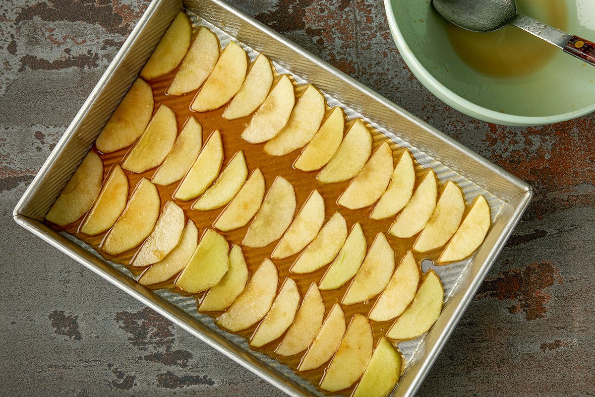 Overhead shot of a rectangular baking pan filled with neatly arranged apple slices on a layer of syrup; placed beside a green bowl containing liquid and a spoon