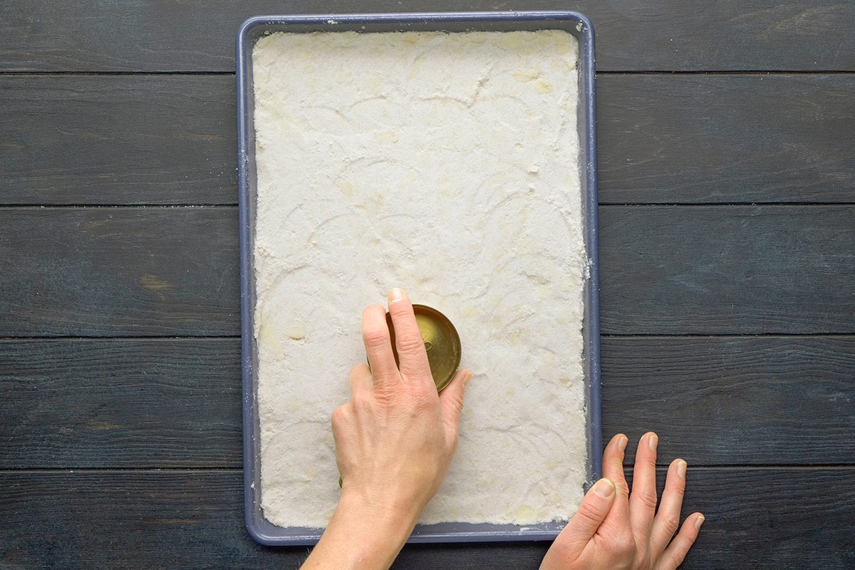 A person presses a round cutter into dough spread evenly in a rectangular baking pan on a dark wooden surface.