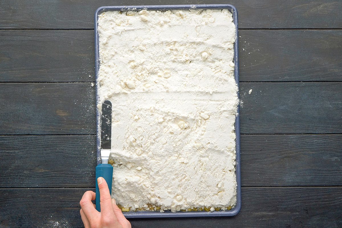A hand holds a spatula, spreading flour evenly across a baking sheet on a dark wooden surface.