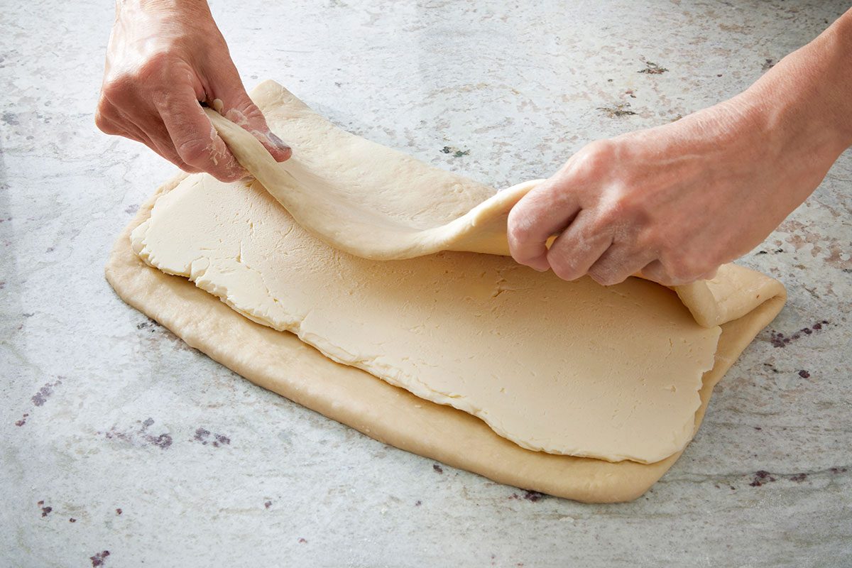 Overhead shot of hands folding a layer of dough over a sheet of butter on a lightly floured surface; demonstrating the lamination process for pastries such as croissants;