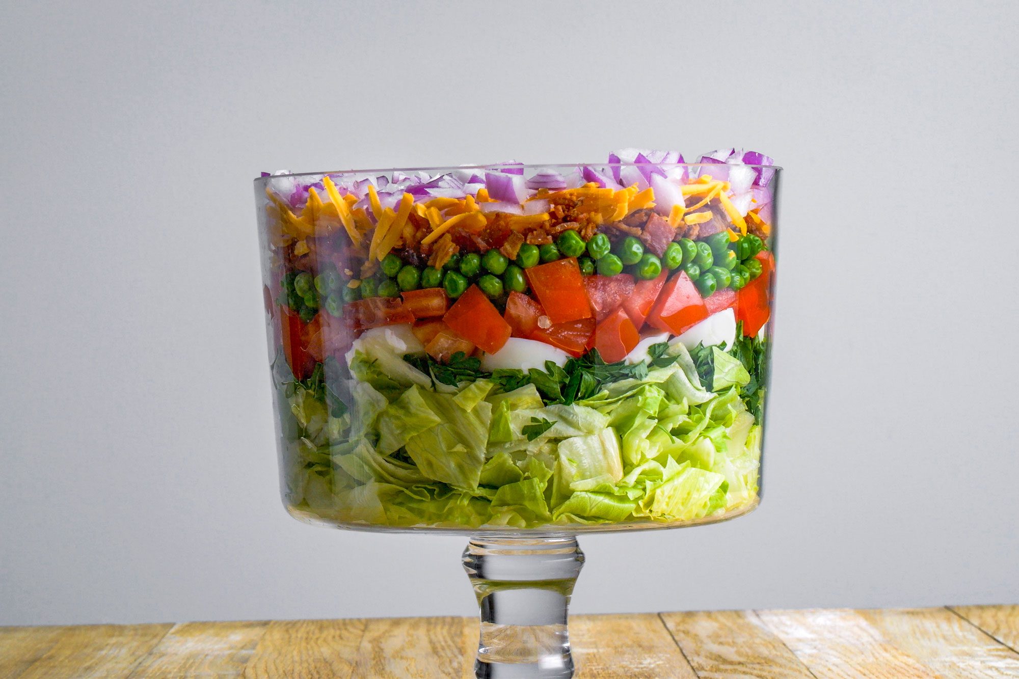 Close shot of a glass trifle bowl filled with layered salad lettuce, spinach, tomatoes, peas, chopped onions, shredded cheese, and bacon displayed on a wooden surface against a plain gray background;