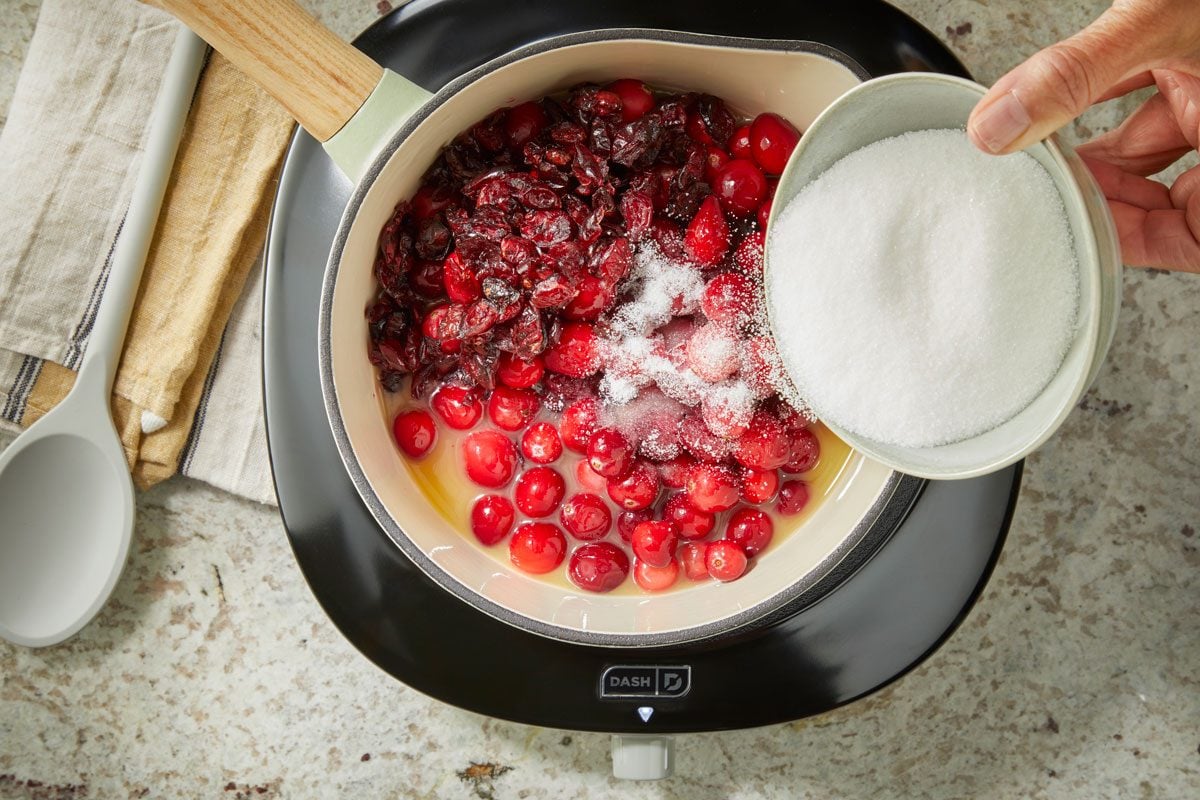 Overhead shot of a hand pouring sugar from a bowl into a saucepan of fresh cranberries and juice on a stovetop, with a spoon and kitchen towels nearby;