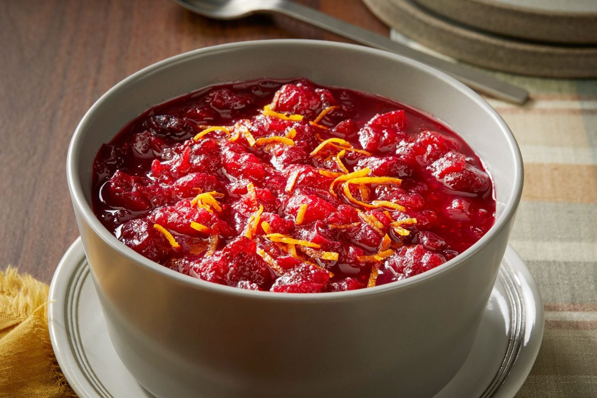 Close view shot of Aperol Spritz Cranberry Sauce served in a bowl, topped with orange zest, and set on a saucer over a checkered tablecloth, with plates and a spoon beside it;
