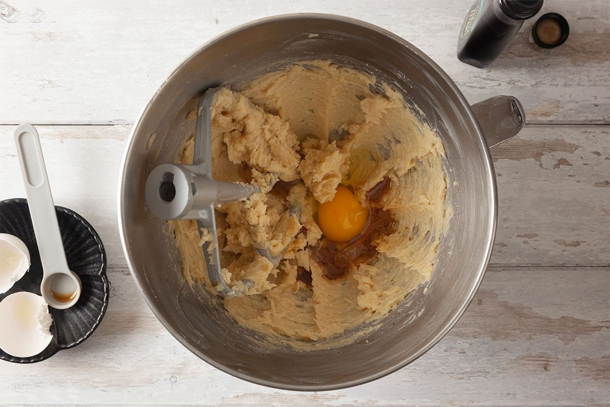 A mixing bowl with creamed butter and sugar, a cracked egg, and vanilla extract about to be mixed. Eggshells and a measuring spoon are nearby on a light wooden surface.