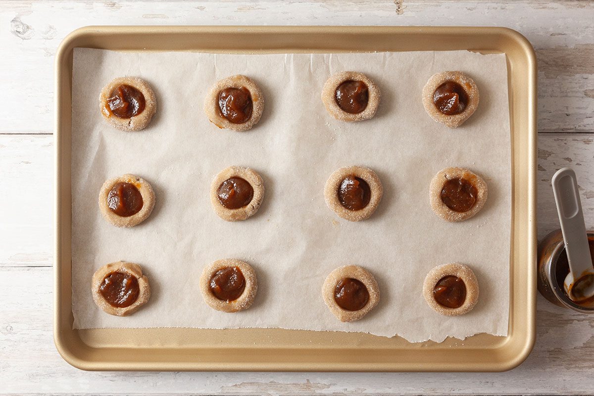 A baking tray lined with parchment paper holds 15 unbaked thumbprint cookies, each filled with caramel or a similar filling. A measuring spoon and small bowl are visible on the side of the tray.