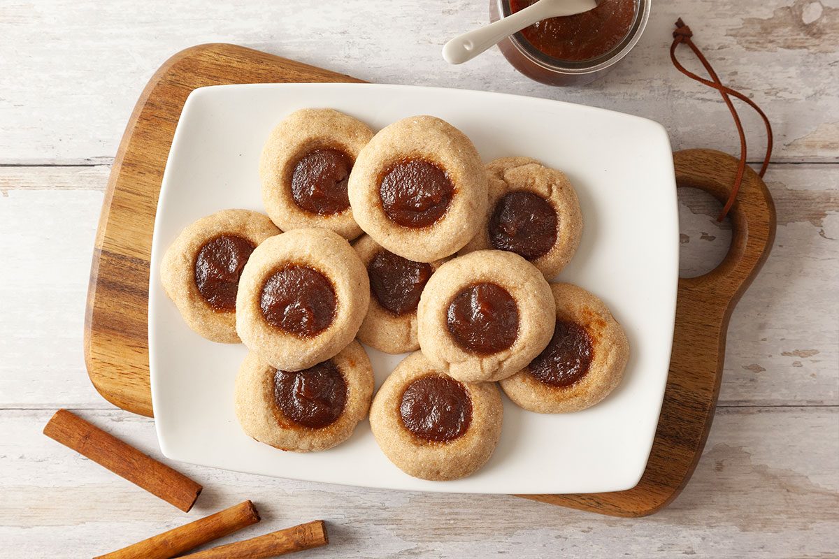 A white plate with round thumbprint cookies filled with brown jam sits on a wooden board. A small jar of jam and two cinnamon sticks are nearby on a light wood surface.