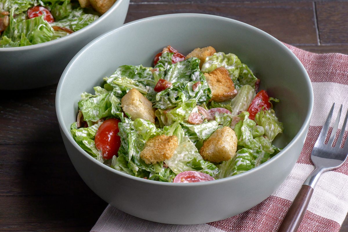 A bowl of fresh salad with lettuce, cherry tomatoes, croutons, and dressing sits on a table next to a fork and a folded striped napkin.