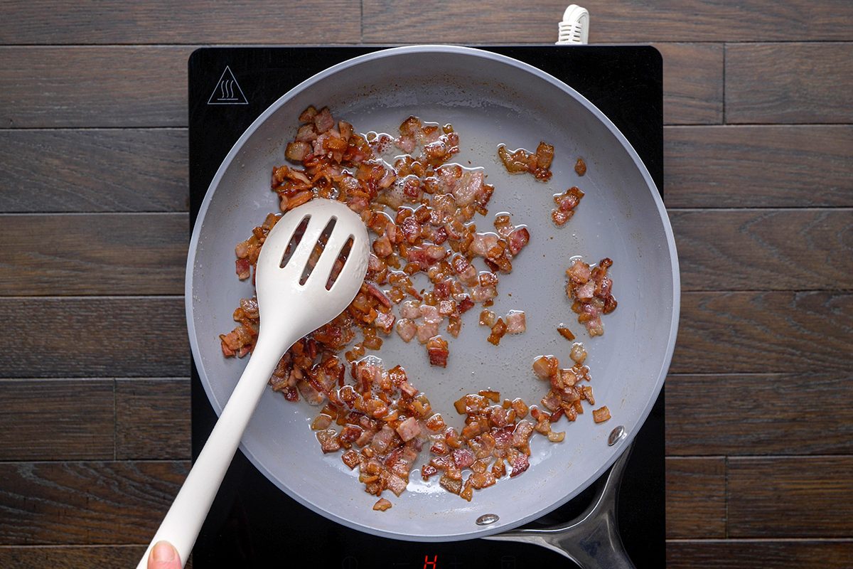 Diced bacon pieces are being cooked in a frying pan on a stovetop, stirred with a white slotted spoon. The bacon is browned and sizzling.